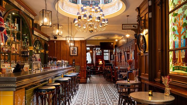 The Victorian interior of the pub with shelves behind the bar filled with bottles, a chandelier,  tables and bar stools