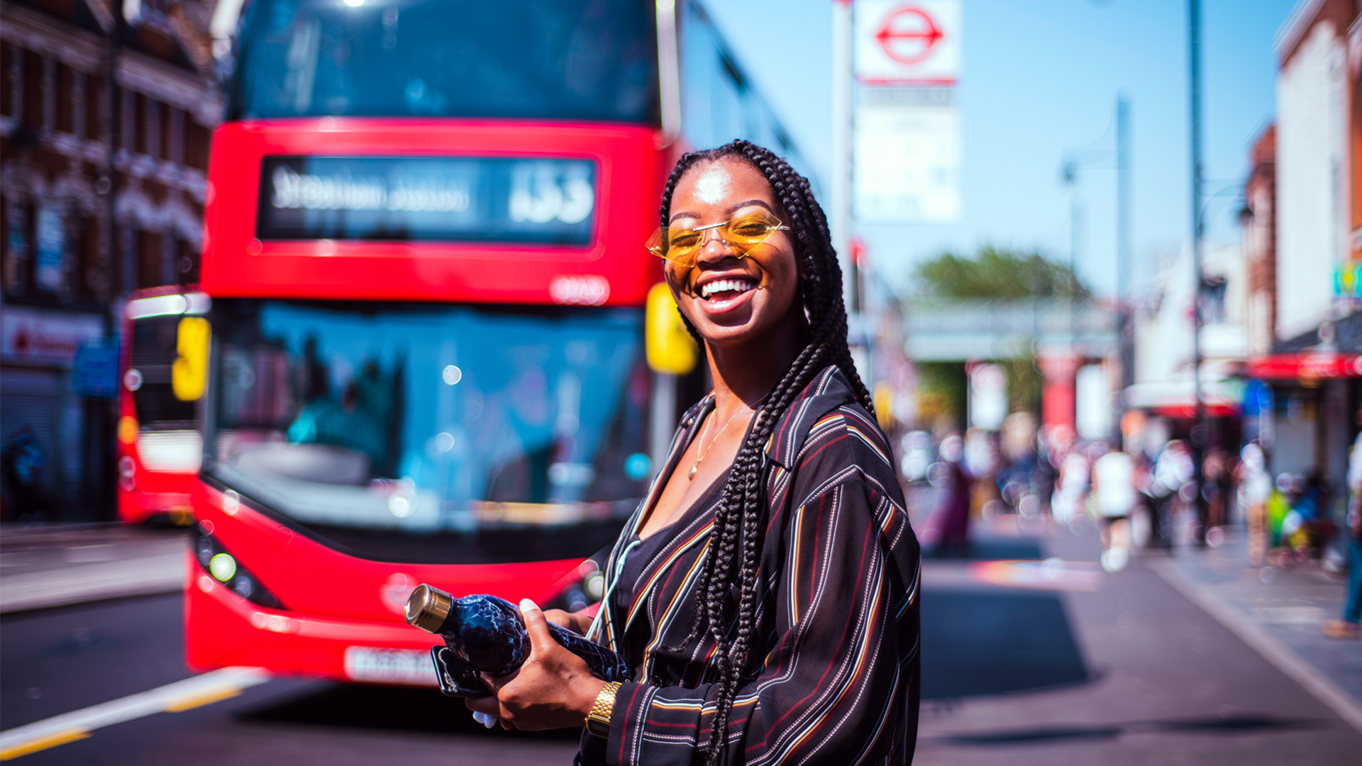 Une jeune femme portant des lunettes de soleil se tient dans les rues de Londres par une journée ensoleillée alors qu'un bus rouge à impérial approche.