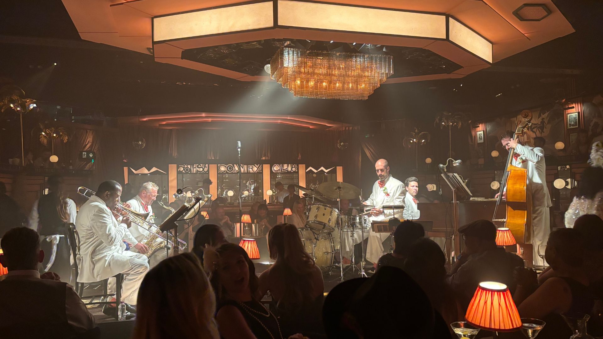 Listen to electric live jazz performances, led by the enigmatic Bell Livingstone. Image courtesy of Visit London. Image courtesy of Visit London. A jazz band performs on stage in a dimly lit, elegant club. The band includes a saxophonist, drummer, and upright bassist, all dressed in white suits with red bow ties. The audience sits at tables lit by small lamps, with a large chandelier hanging above the stage, enhancing the sophisticated ambience.