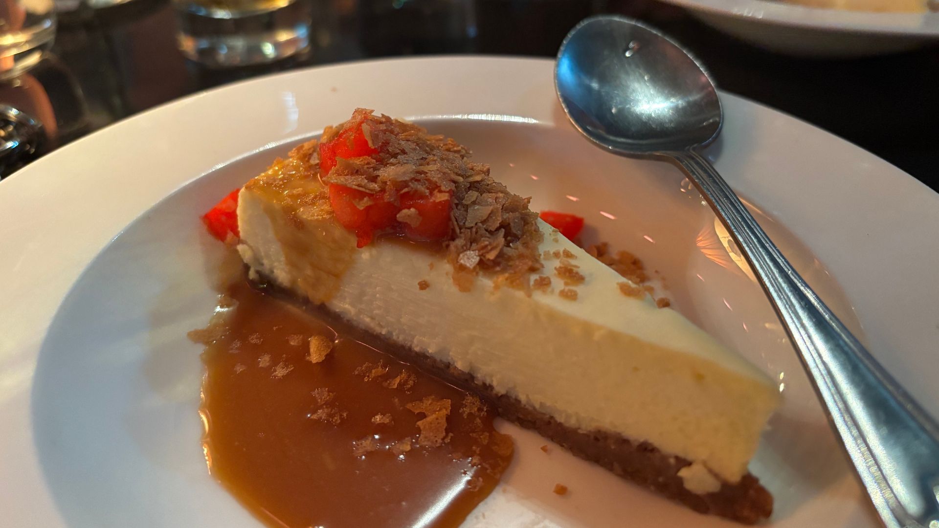 A slice of cheesecake topped with chopped strawberries and a crumbly topping, served on a white plate with caramel sauce. A spoon rests on the edge of the plate.