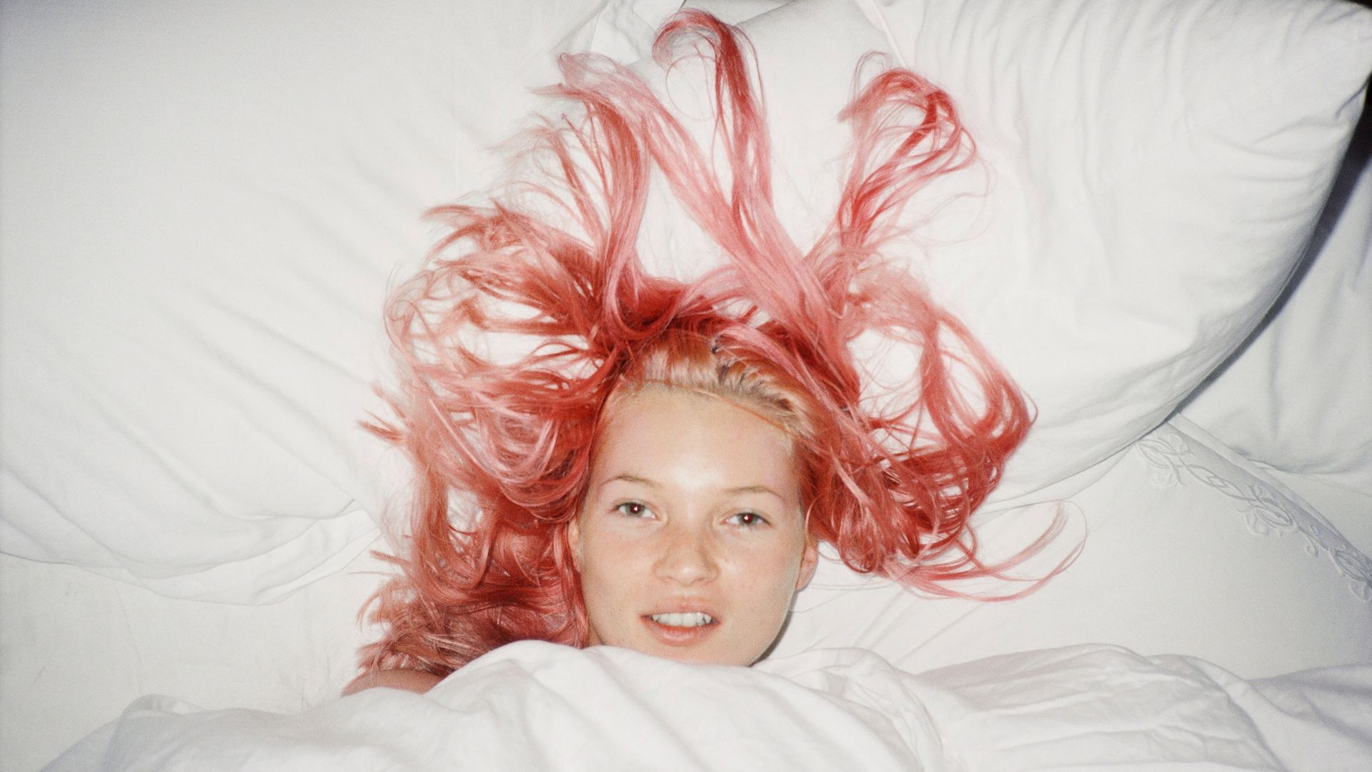 A photograph of a woman lying in bed with her long pink hair spread out across white pillows and bedding.