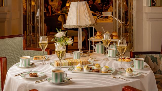 Round table set for afternoon tea at Claridge’s with sandwiches, cakes, scones, tea cups and glasses of sparkling wine in a luxurious dining room.