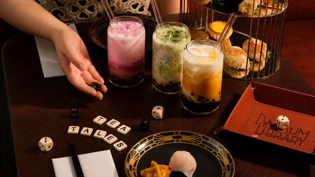 Three bubble tea drinks served alongside dim sum, pastries and board games on a dark wood table at Dim Sum Library.