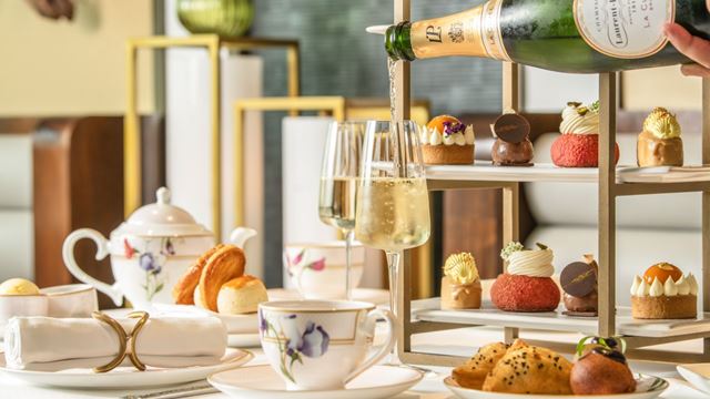 Tiered afternoon tea stand displaying assorted desserts and savouries, with Champagne, tea service and scones arranged on a white tablecloth in an elegant restaurant setting.