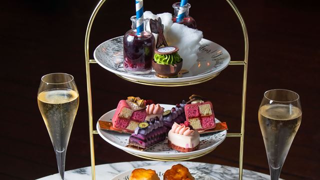 Three-tier afternoon tea stand displaying Alice in Wonderland-themed desserts, including colourful cakes, a pocket watch-inspired sweet and bottle-shaped treats, served with glasses of sparkling wine on a marble table.