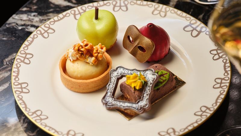 Selection of theatrical-themed afternoon tea pastries on a decorative plate, including an apple-shaped dessert, a red mousse topped with a gold theatre mask, a caramel tart and a chocolate cake decorated with an ornate picture frame.