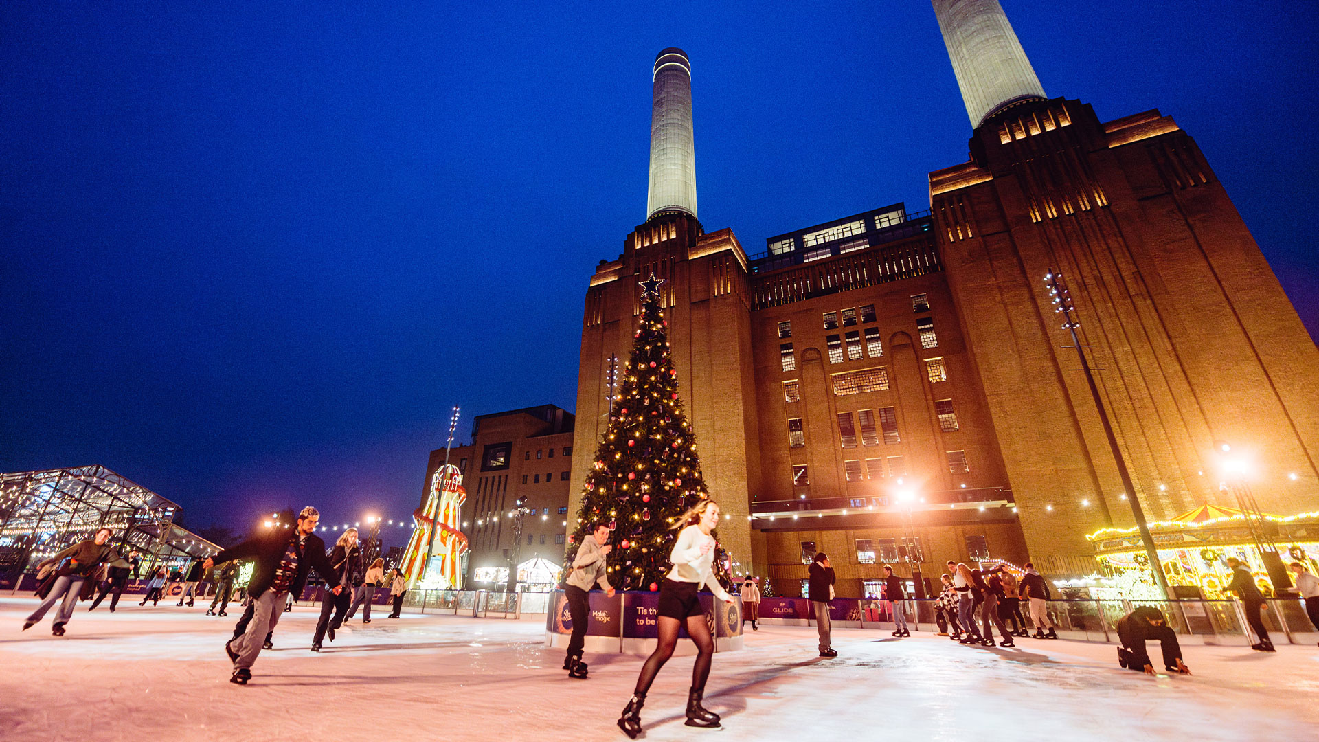 Skaters glide along the ice at Glide at Battersea Station, an ice rink located in front of the power station and featuring a Christmas tree on the ice.