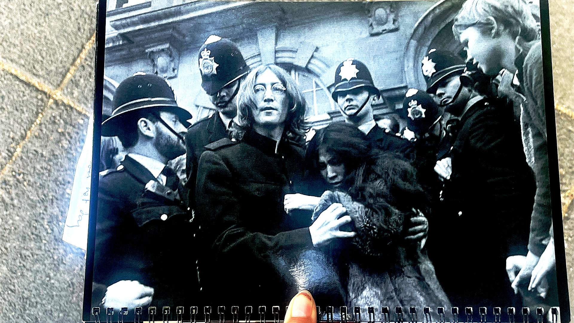 Someone is holding up a black-and-white photograph of John Lennon and Yoko Ono, surrounded by police officers, on the doorstep of their flat on Montagu Square.