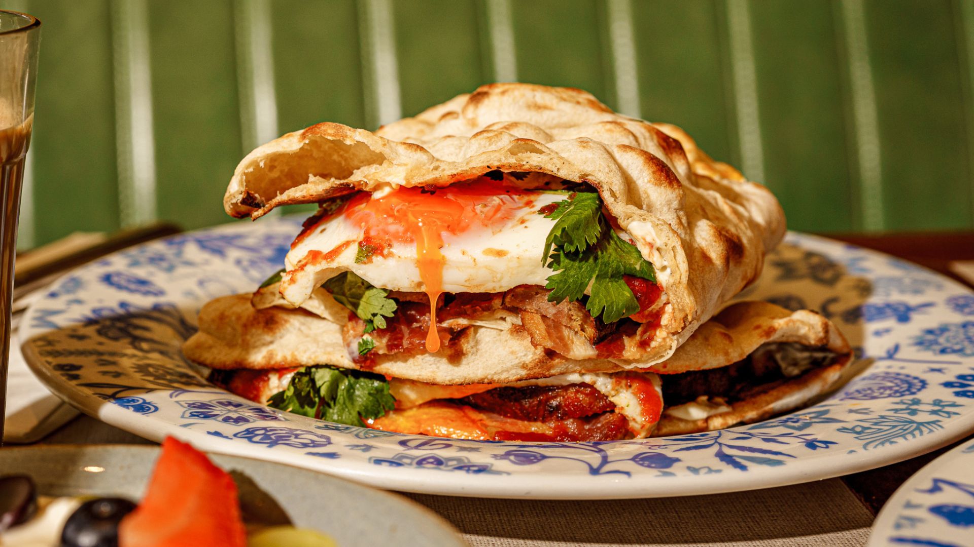 a naan bread stuffed with bacon, sausage and a runny yolked egg, presented on a small flowery white and blue plate