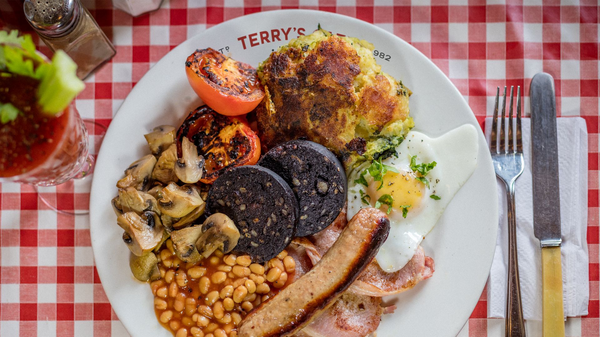 Enjoy a traditional English breakfast at the iconic Terry's Cafe in London. Image courtesy of Terry's Cafe. high angle photo of a white plate of beans, sausage, egg, bacon, mushrooms, tomatoes, bubble and squeak and black pudding, on a chequered red and white tablecloth
