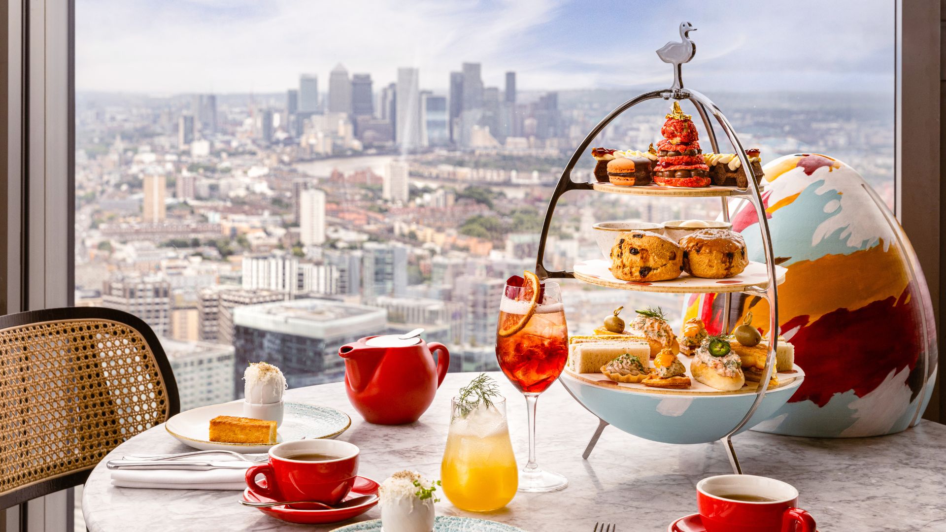 Tiered afternoon tea stand with pastries, scones and sandwiches beside tea and cocktails, set against a panoramic view of London’s skyline.