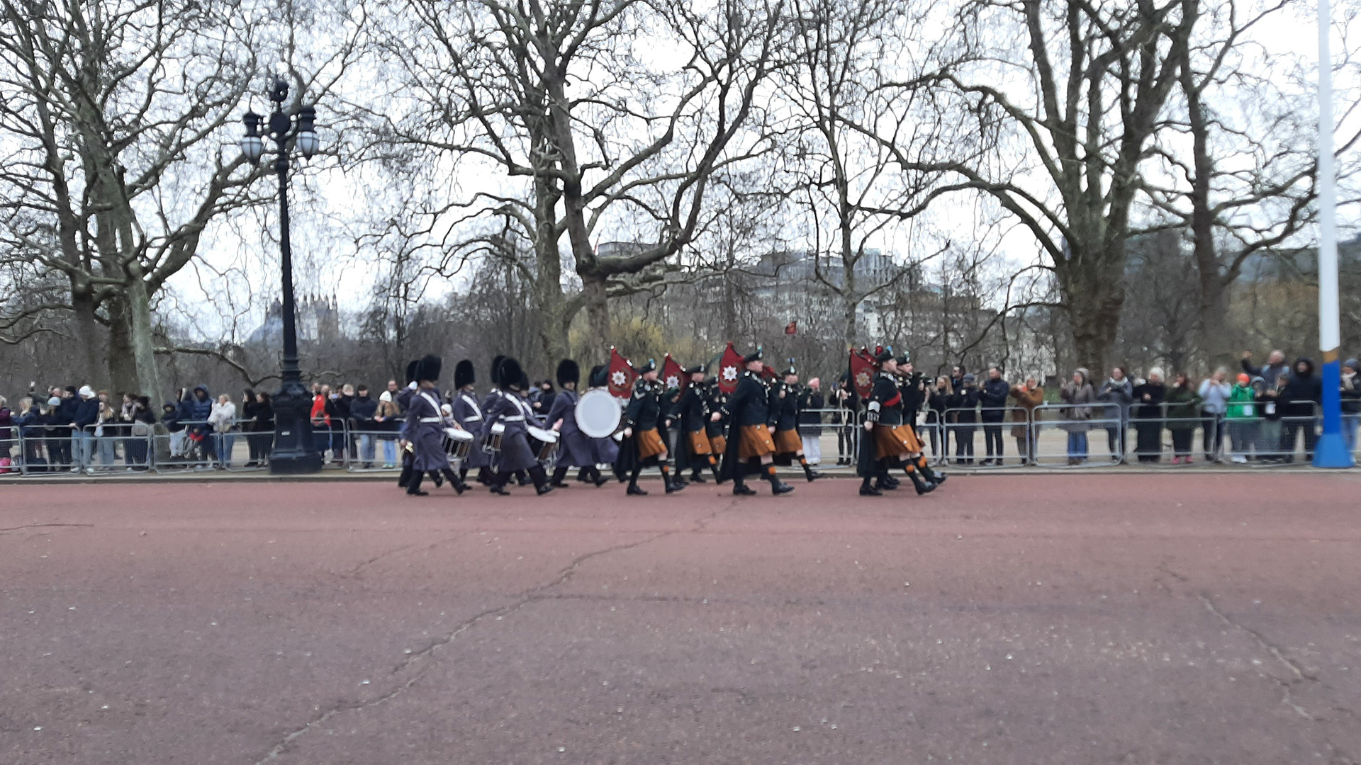 Foot soldiers in blue/grey long winter greatcoats wearing a bearskin while marching with drums and soldiers marching in front who wear yellow quilts and berets