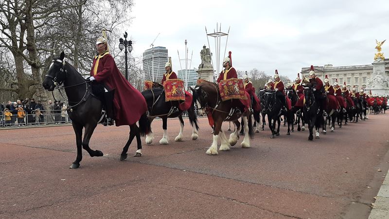 Mounted soldiers with instruments and red long cloaks and helmets with blumes march along The Mall away from Buckingham Palace.