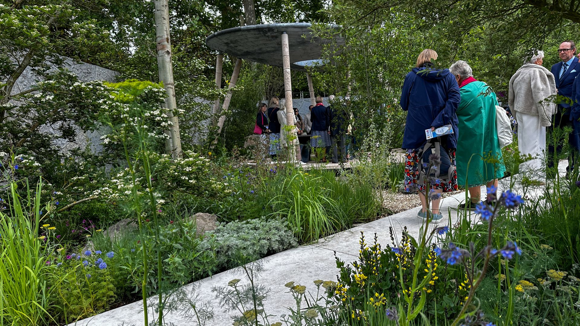Visitors walk through the Eden Project Garden past mixed edible and ornamental planting towards a pavilion with a curved roof supported by angled legs, designed using sustainable materials.