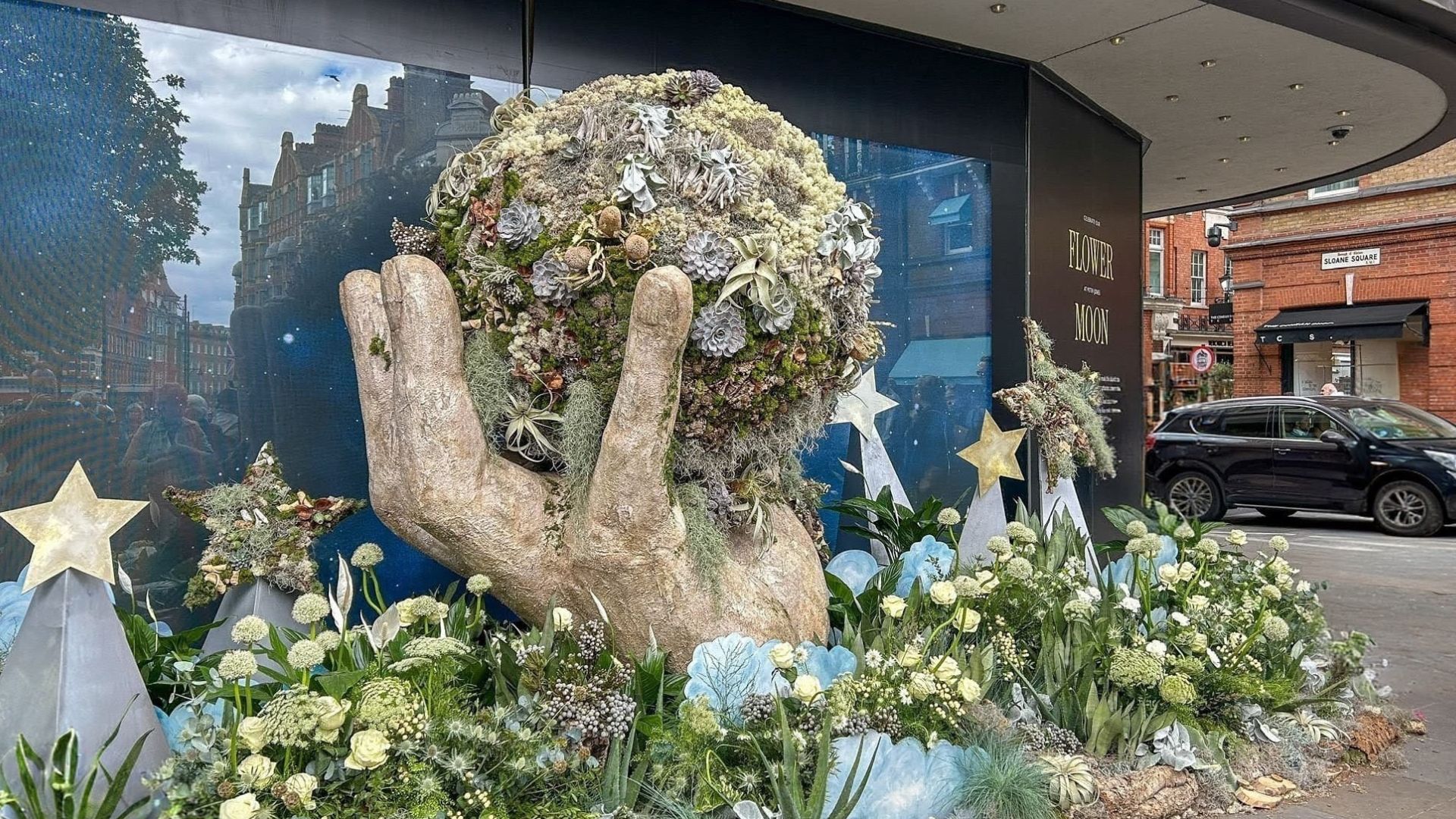Large sculpted hand holding a plant-covered globe, surrounded by white flowers and star shapes outside a Sloane Square shop display