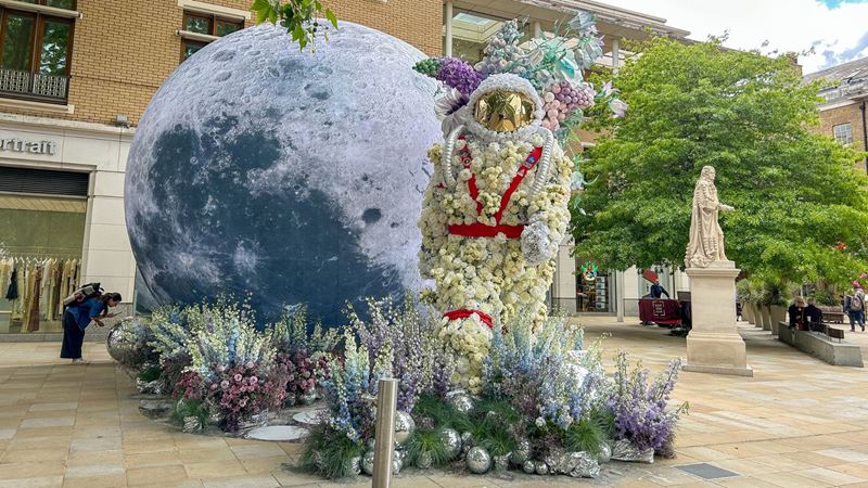 Flower-covered astronaut figure beside a giant moon installation, surrounded by pastel flowers in an outdoor courtyard setting