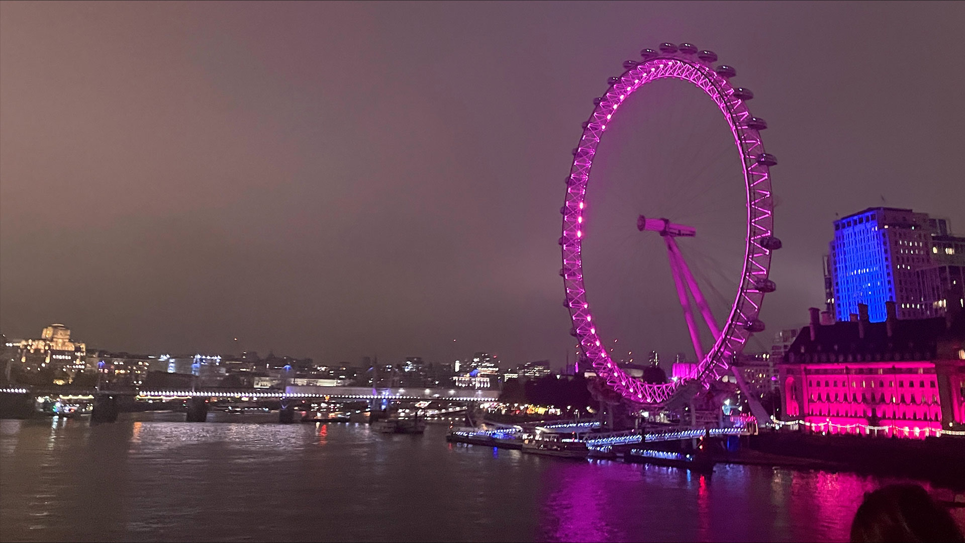 The London Eye lit up in pink and reflecting onto the river Thames at night