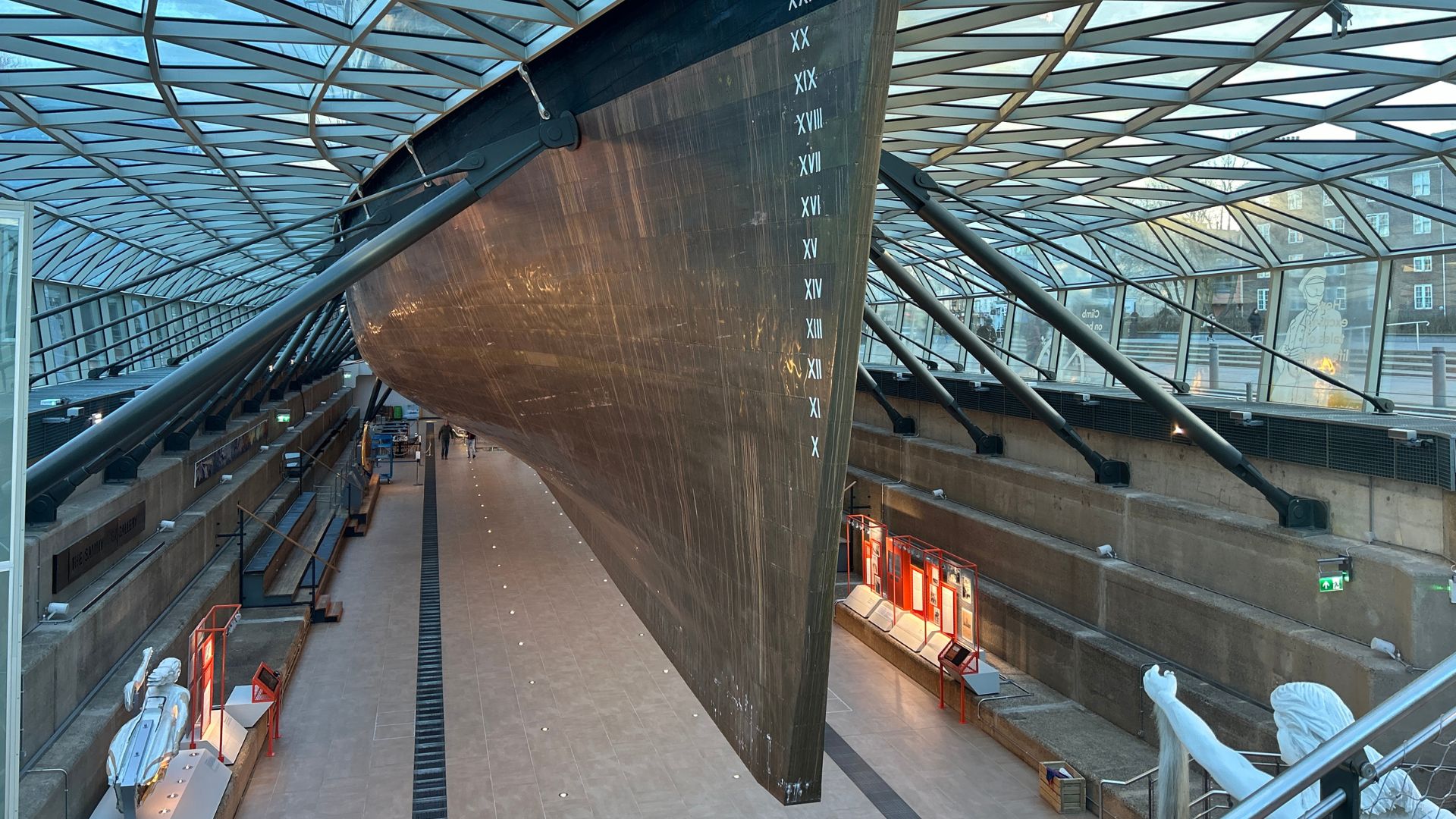 Tuck into traditional afternoon tea, served beneath the iconic copper clad hull at the Cutty Sark. © Visit London/Hannah Cornish a large ship held up with structuring and a glass roof above, with people walking beneath the ship museum