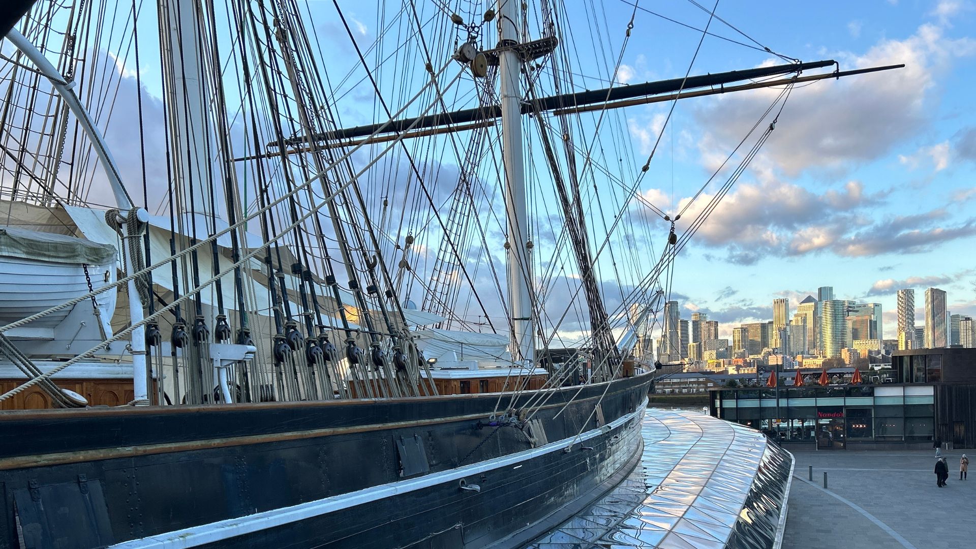 Enjoy panoramic views of London's iconic skyline from the deck of the iconic Cutty Sark. © Visit London/Hannah Cornish a side view of the Cutty Sark boat, with the skyscrapers and city buildings in the background, with blue cloudy skies
