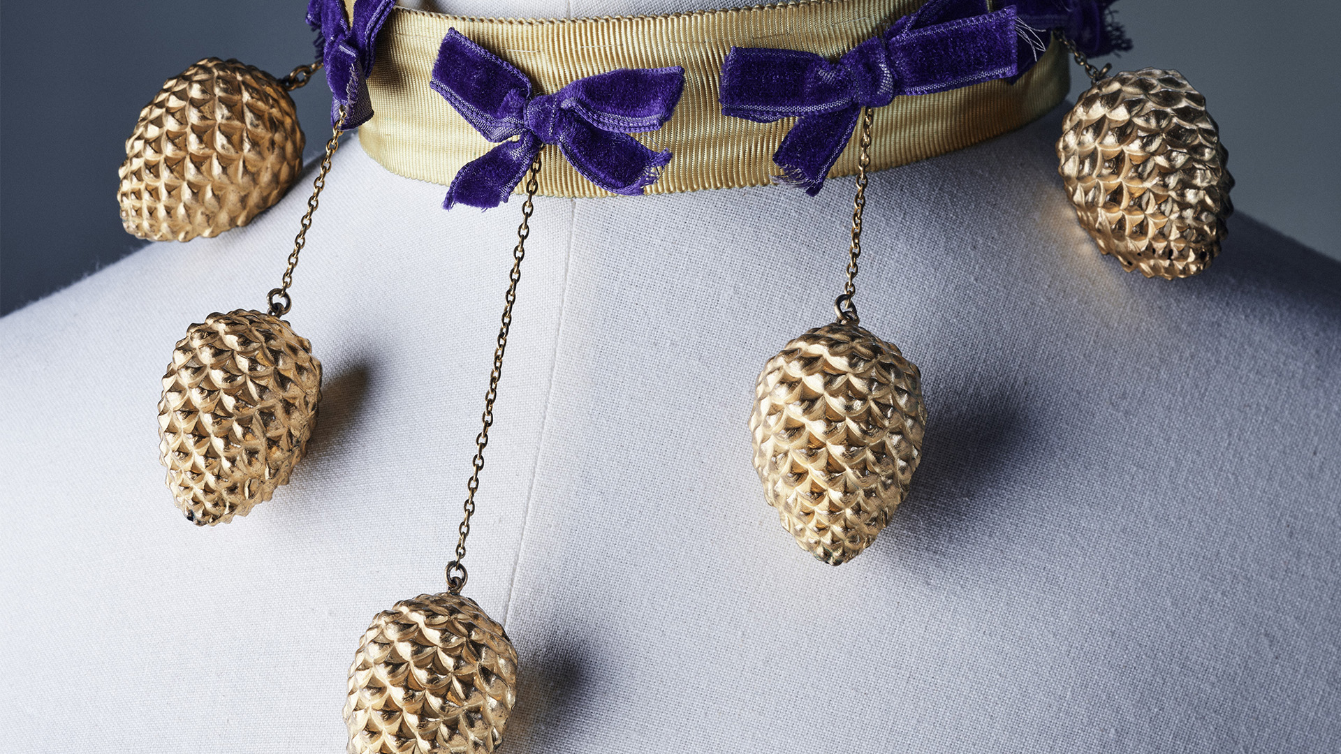 A choker necklace with dangling golden pine cones attached to a golden chocker necklace with purple bows.