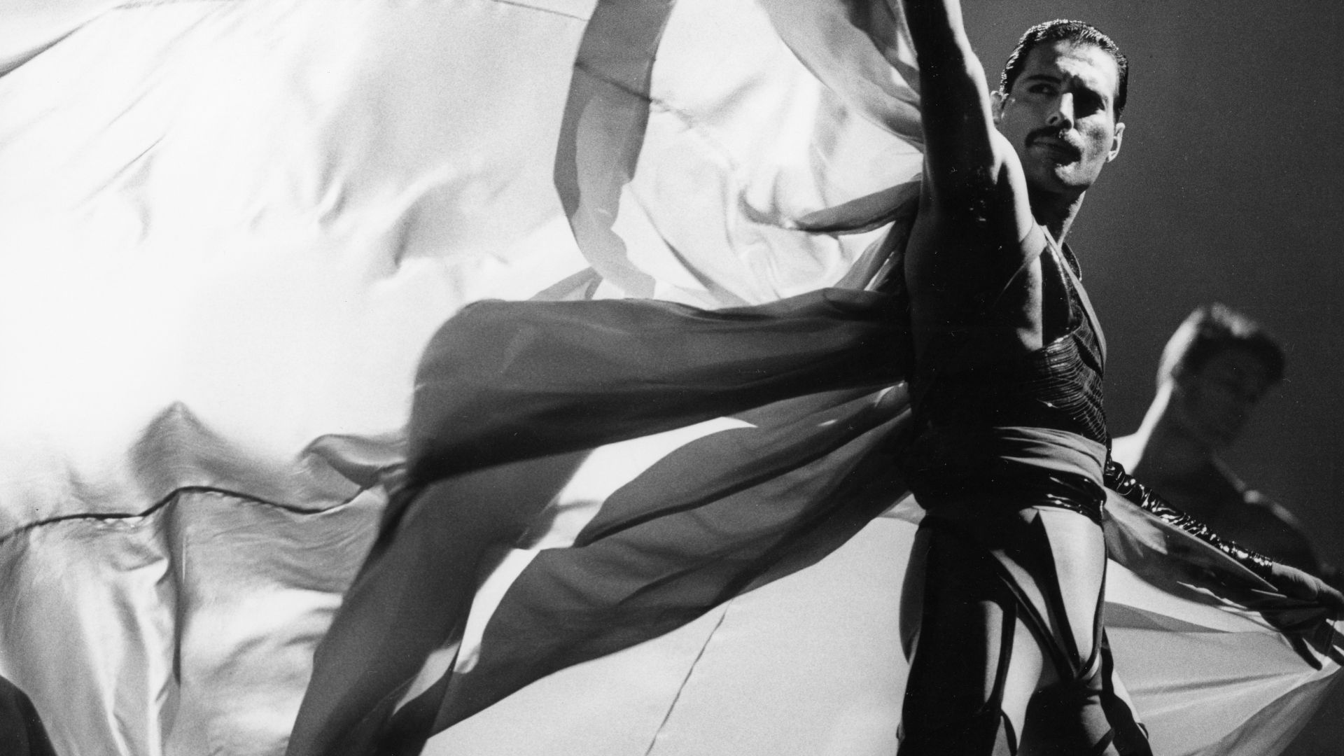 See the iconic outfit worn by Freddie Mercury in the Made in Heaven music video. © Simon Fowler/V&A a black and white photo with a man wearing a dark, tight fitting suit and a large, light cape bellowing in the wind behind him as he looks just past the cameras