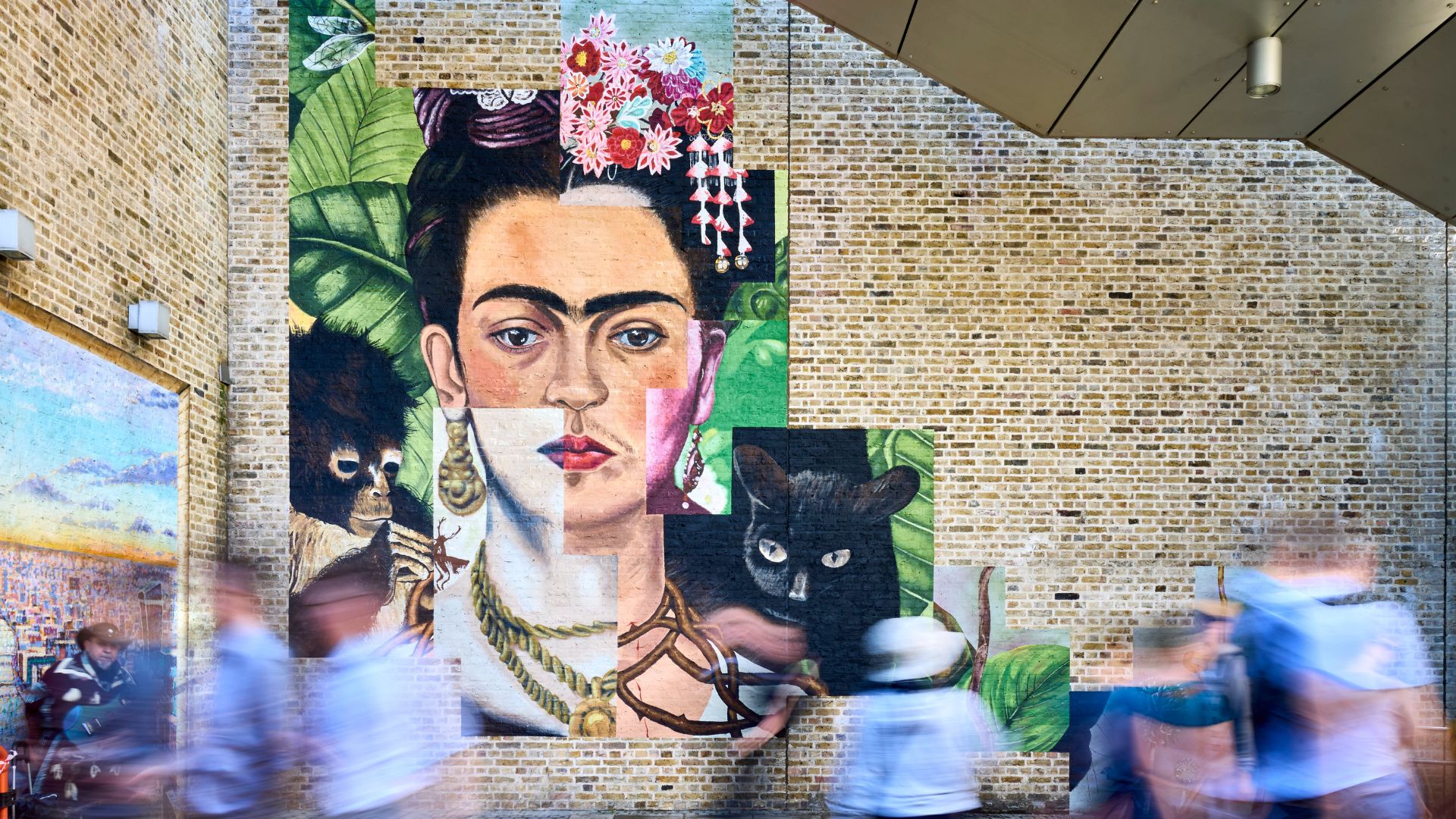 Visitors walk past a large Frida Kahlo mural inside Tate Modern in London, featuring a collage-style portrait inspired by the artist's iconic self-portraits.