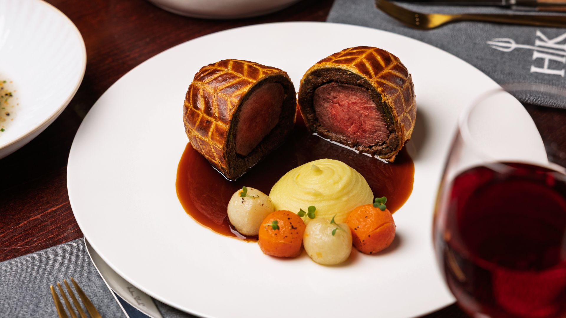 The signature Beef Wellington at Gordon Ramsay Hell's Kitchen London - perfected pastry, tender beef and rich, indulgent flavour. Image courtesy of Gordon Ramsay Hell's Kitchen London. Beef Wellington cut in half on a white plate, served with mashed potato, glazed vegetables and red wine sauce.