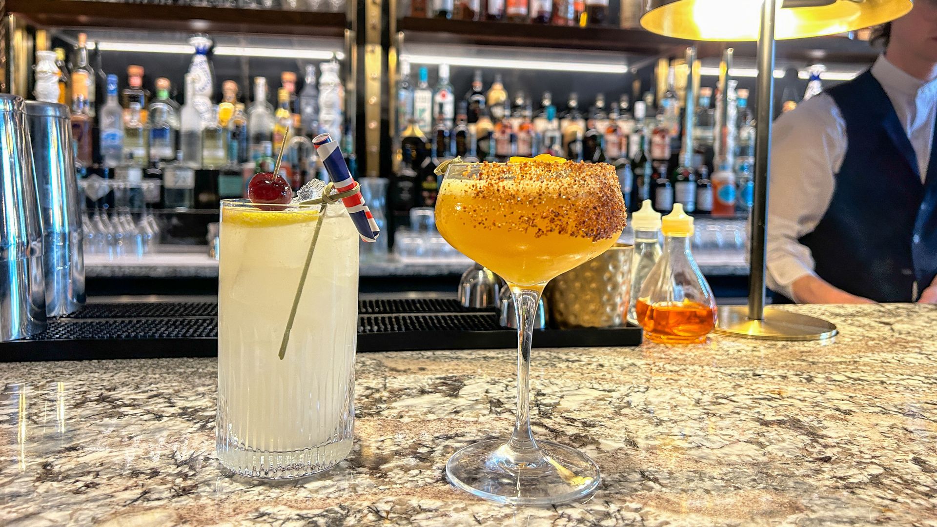 A close up shot of two cocktails on a marble top bar surface, with a smartly dressed barman in the background 