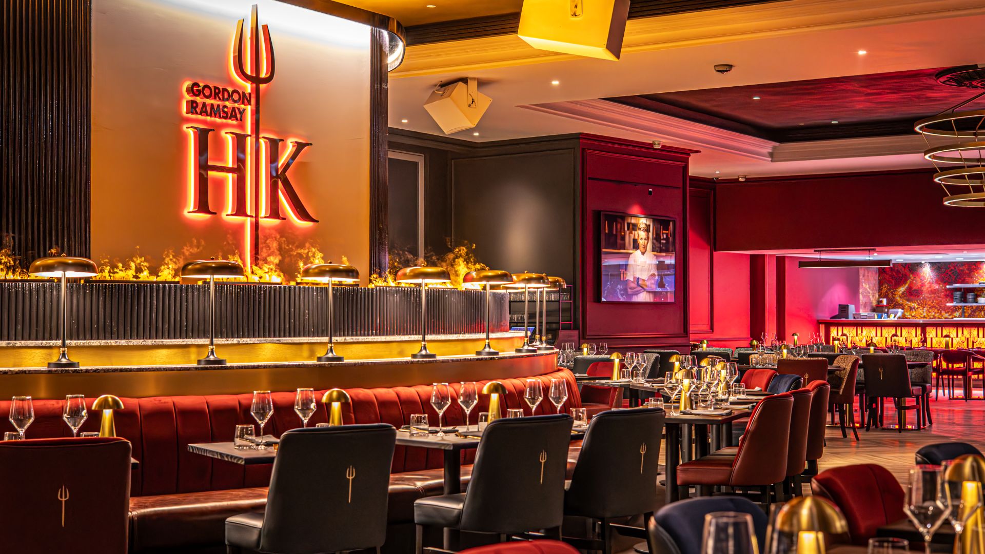 Bold flames and iconic design await – the drama of Hell's Kitchen TV series comes alive. Image courtesy of Gordon Ramsay Hell's Kitchen London. Gordon Ramsay Hell's Kitchen London restaurant interior with red seating, gold lighting, flame features and set dining tables.