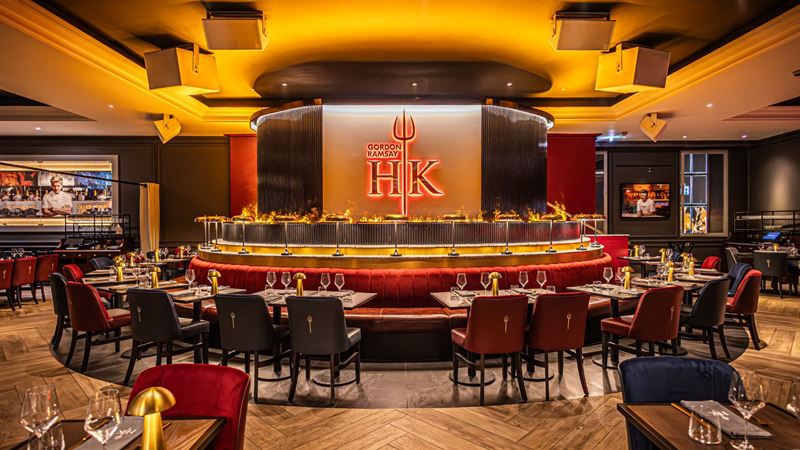 Wide angle view of Gordon Ramsay Hell's Kitchen London dining room with circular central bar, red and blue seating areas and warm, flame lighting.