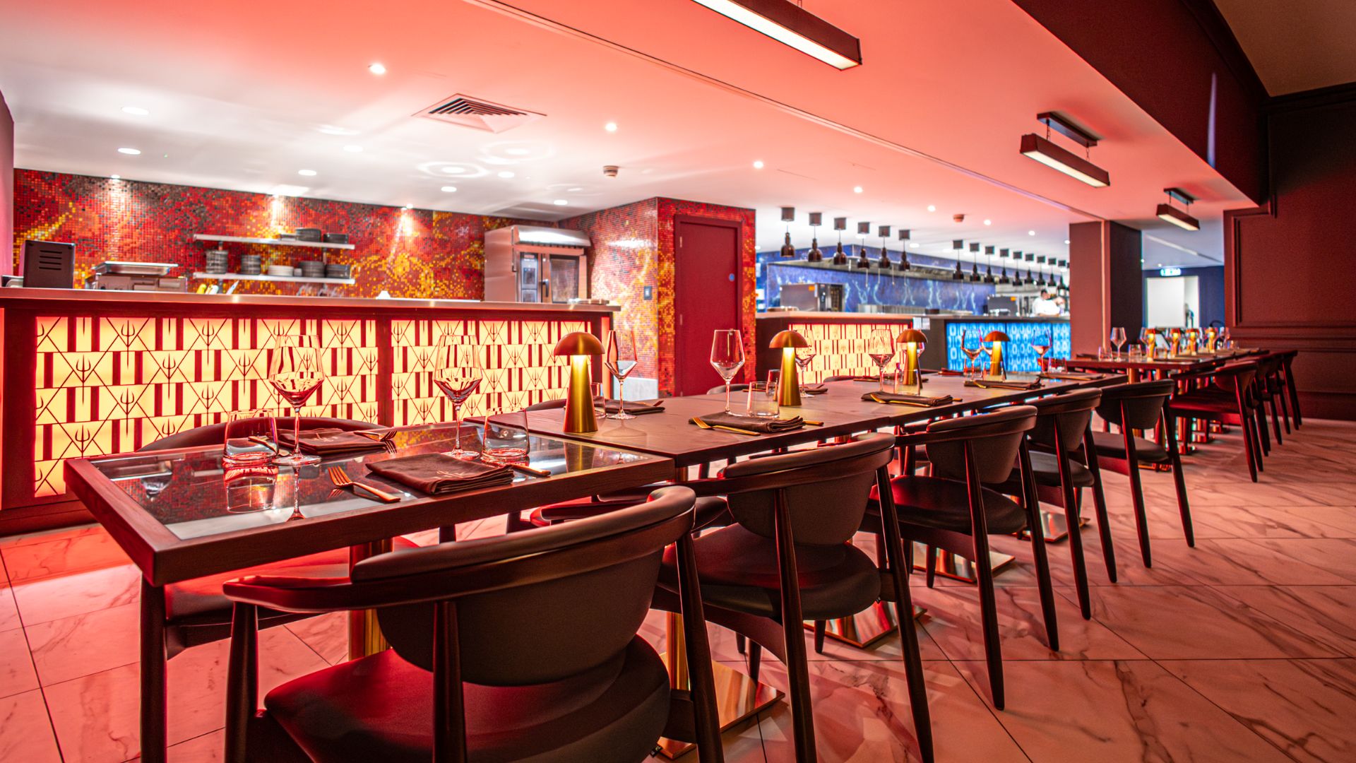 See the Red Kitchen and Blue Kitchen in action, setting the mood for an unforgettable experience and tantalising plates. Image courtesy of Gordon Ramsay Hell's Kitchen London. Restaurant dining area with red lighting, long tables set with wine glasses and illuminated kitchen counters.