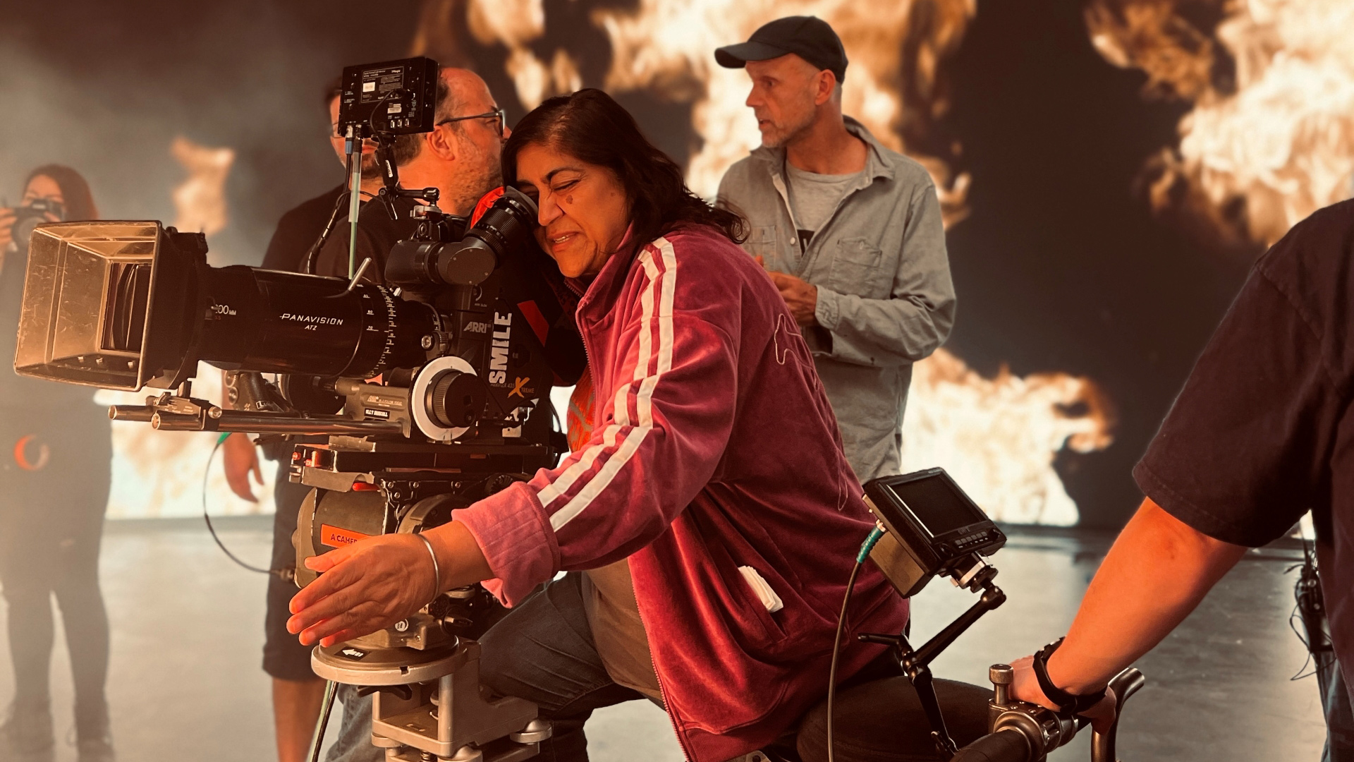 A photo of director Gurinder Chadha looking into a video camera on set with members of the production team in the background