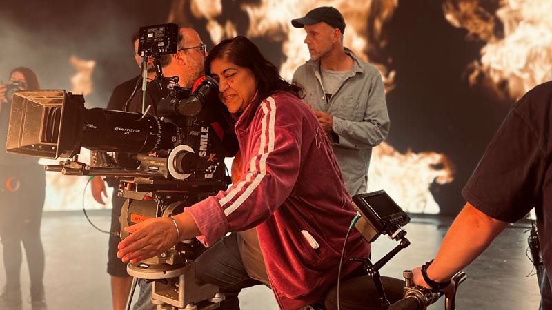 A photo of director Gurinder Chadha looking into a video camera on set with members of the production team in the background