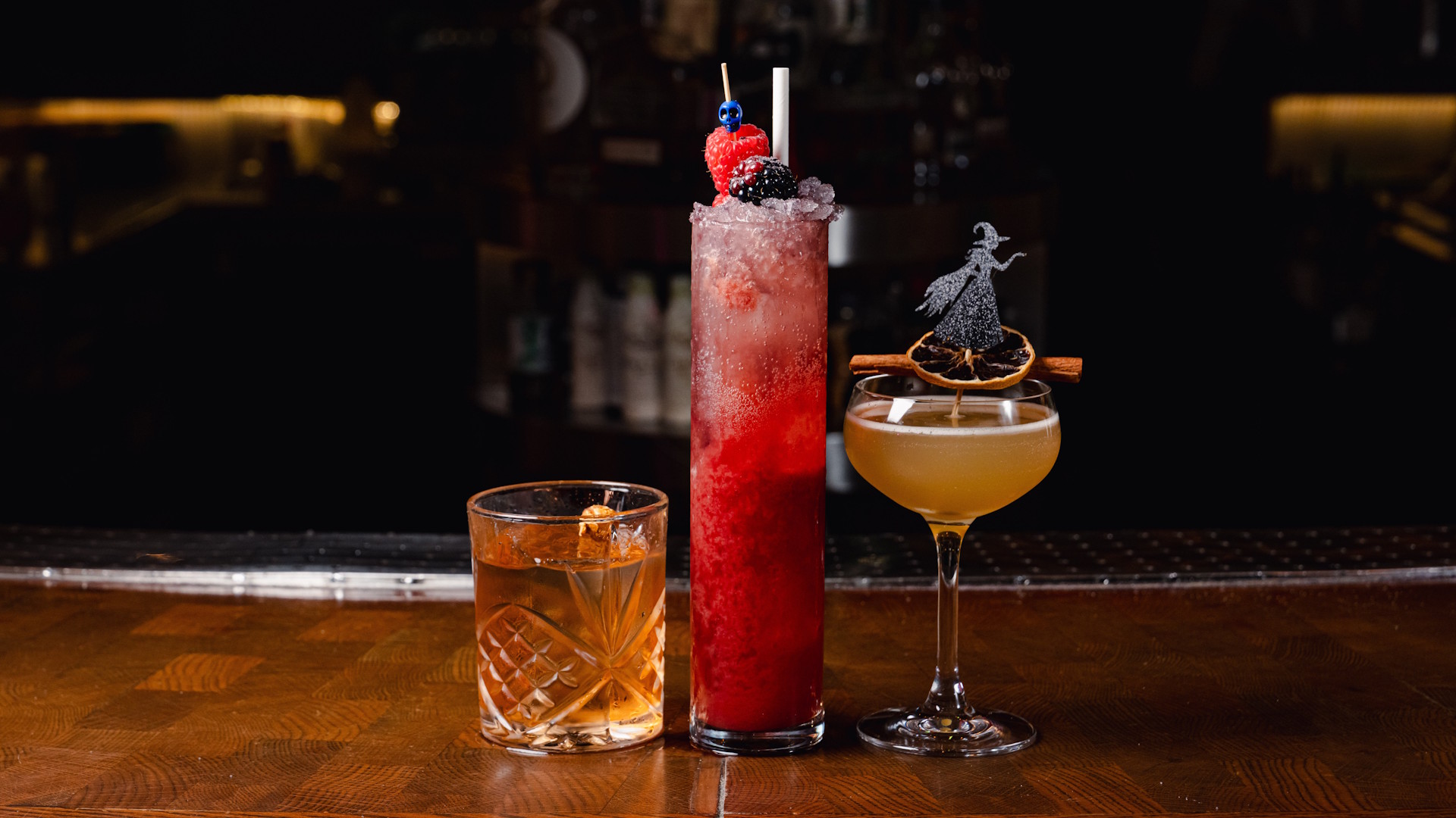 Three orange and red cocktails on a table, including one with a witch on a broomstick balancing on top of the glass.