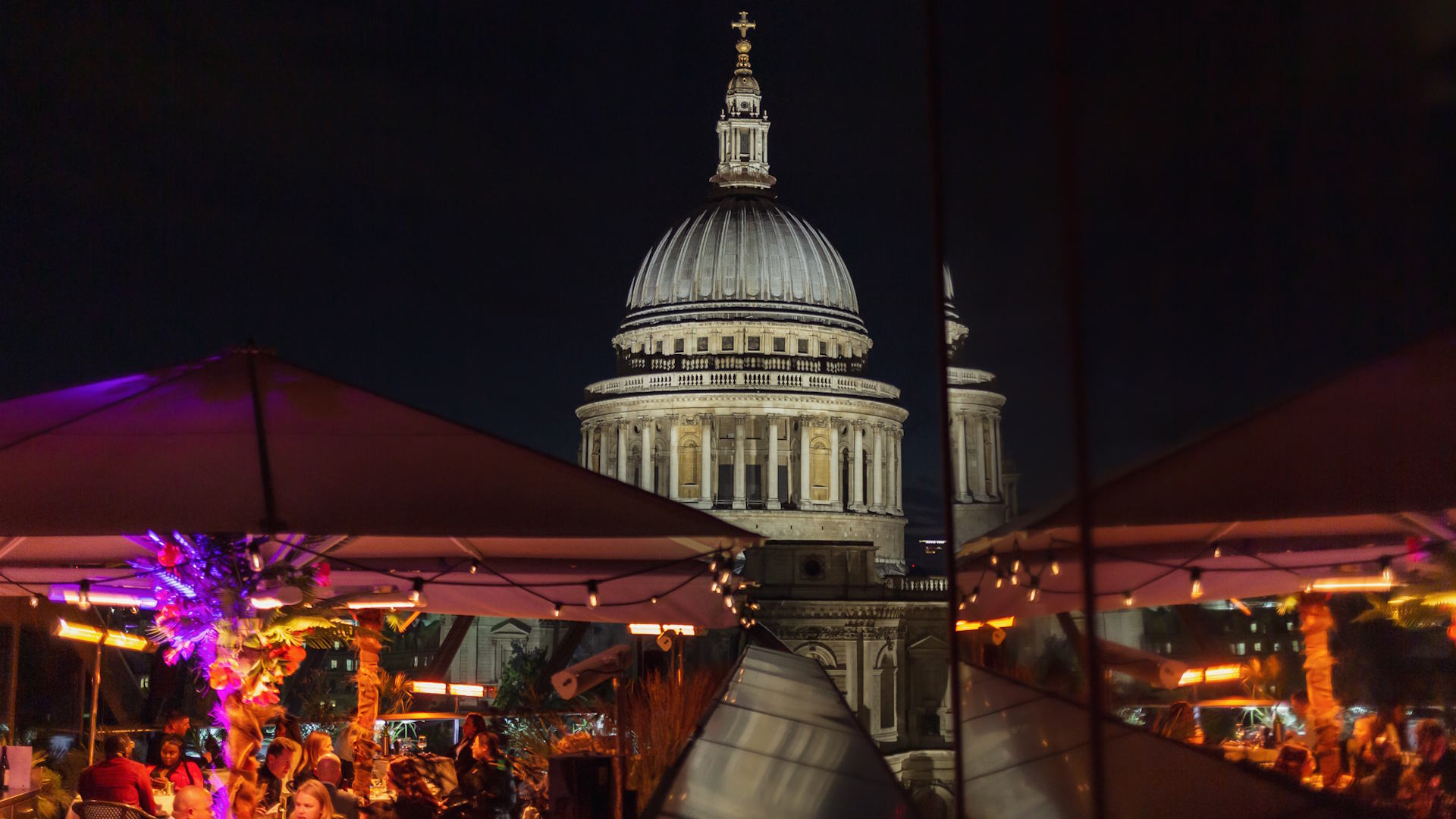 Join Madison Lates' special La Femme Fatale Halloween Party overlooking St Paul's Cathedral. Image courtesy of The Evolv Collective. The rooftop at Madison with orange and purple lighting under parasols where diners sit around tables, and St Paul's Cathedral in the background.