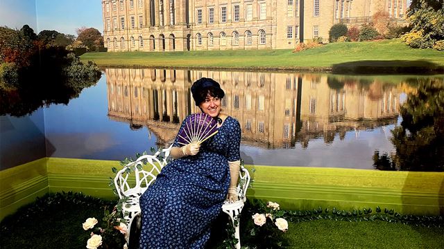 A woman wearing a navy blue Regency-style dress sits on a bench, holding a fan, with a manor by a lake in the background.