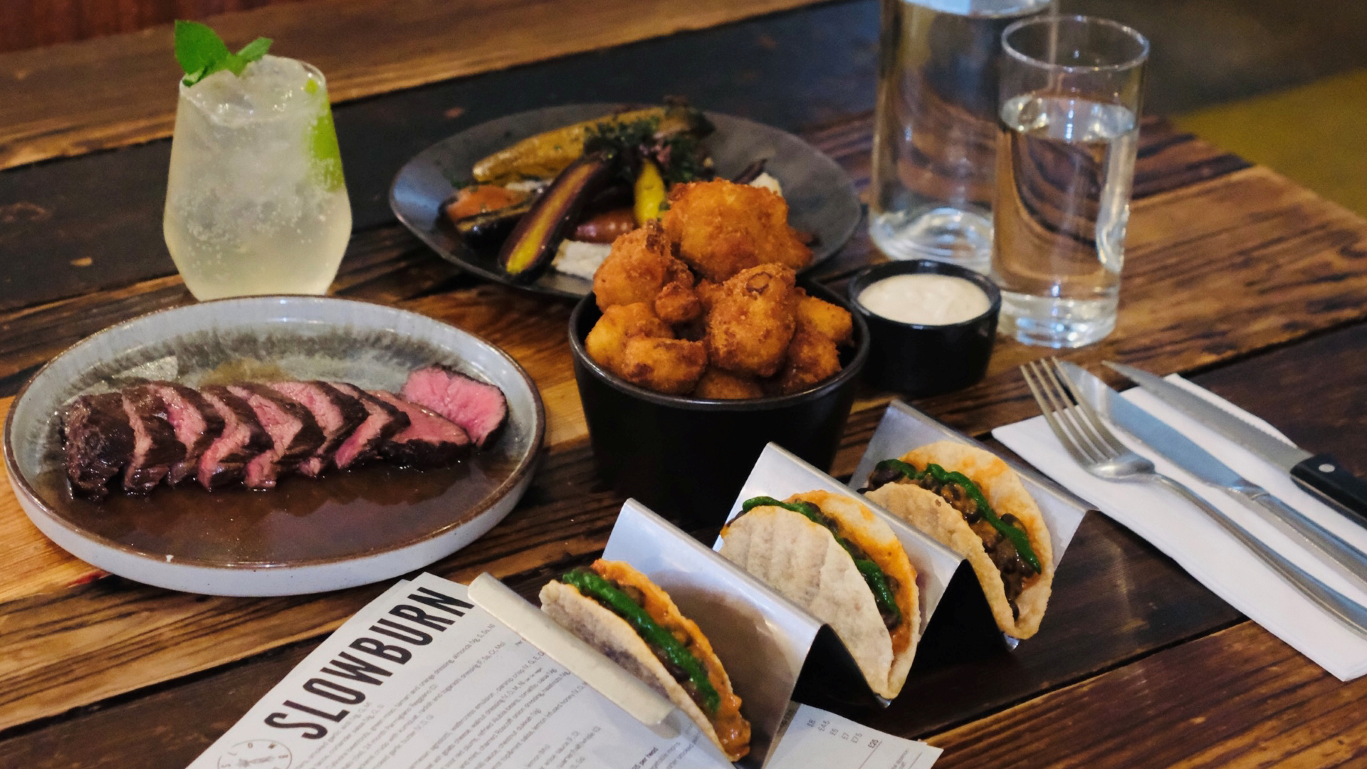 A photo of a table of food at Slowburn, featuring a thinly sliced pink steak on a plate, three tacos, vegetables, and a deep fried side with water, a cocktail and the menu also on the table