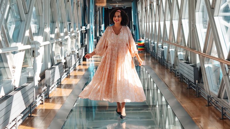 Glass Floors at Tower Bridge. Credit: Jou Jou Travels. Image courtesy of Jou Jou Travels. A photo of Jou Jou Travels travel influencer walking through the glass floor walkway beneath Tower Bridge in a long pink flowing dress with a smile on her face