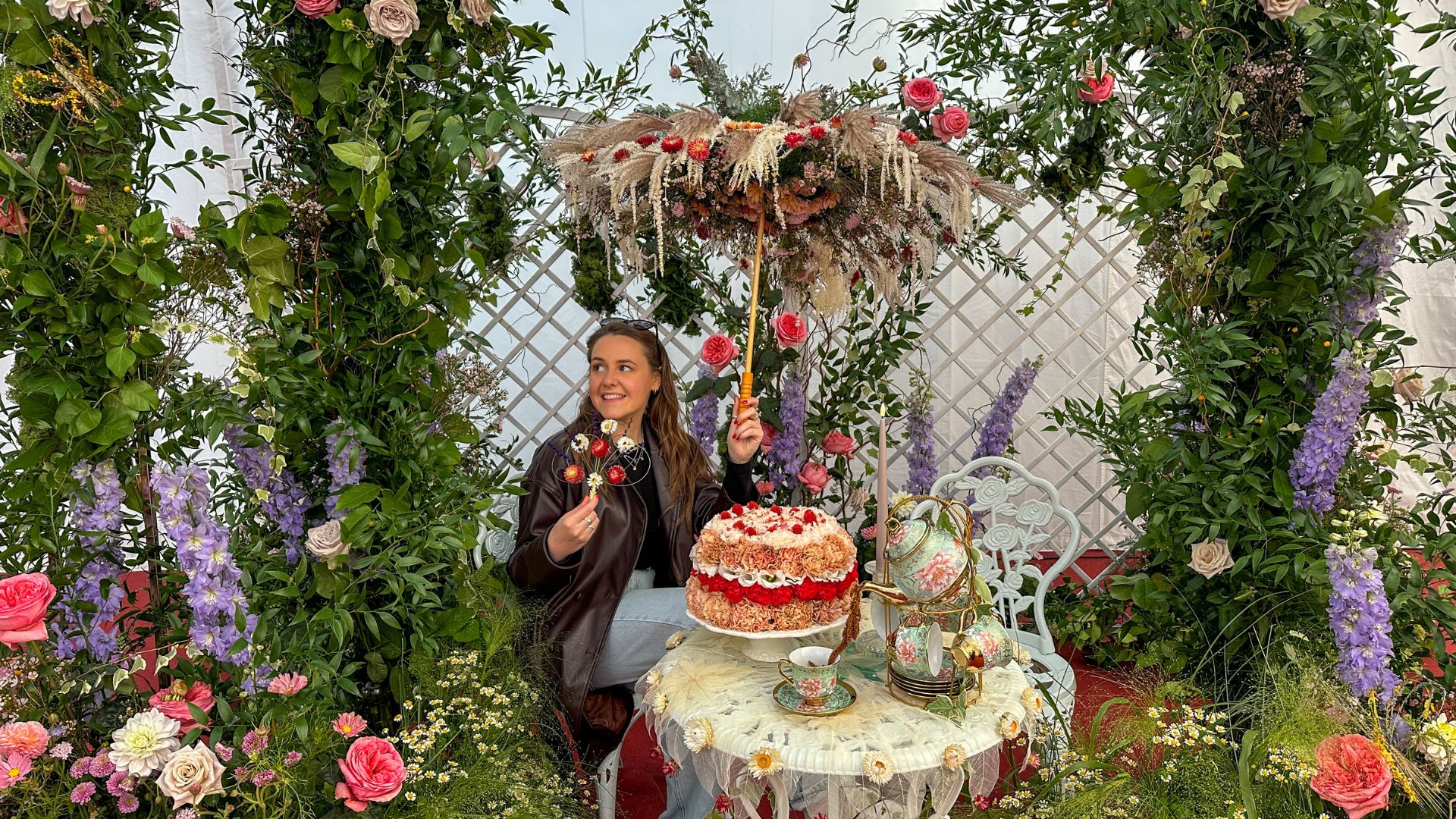 Snap a photo at the interactive floral installations at the Downton Abbey exhibition at Kew Gardens. Photo Credit: Kirstine Spicer. A girl in a brown coat sits next to a floral arrangement of an afternoon tea at the Downton Abbey exhibition at Kew Gardens.