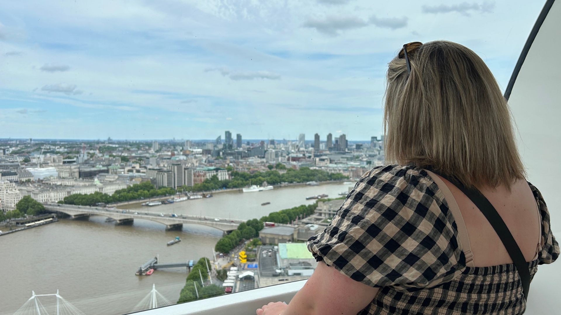 Person looking out from a London Eye capsule over the River Thames, with bridges, riverside buildings and the London skyline visible beneath a cloudy sky.
