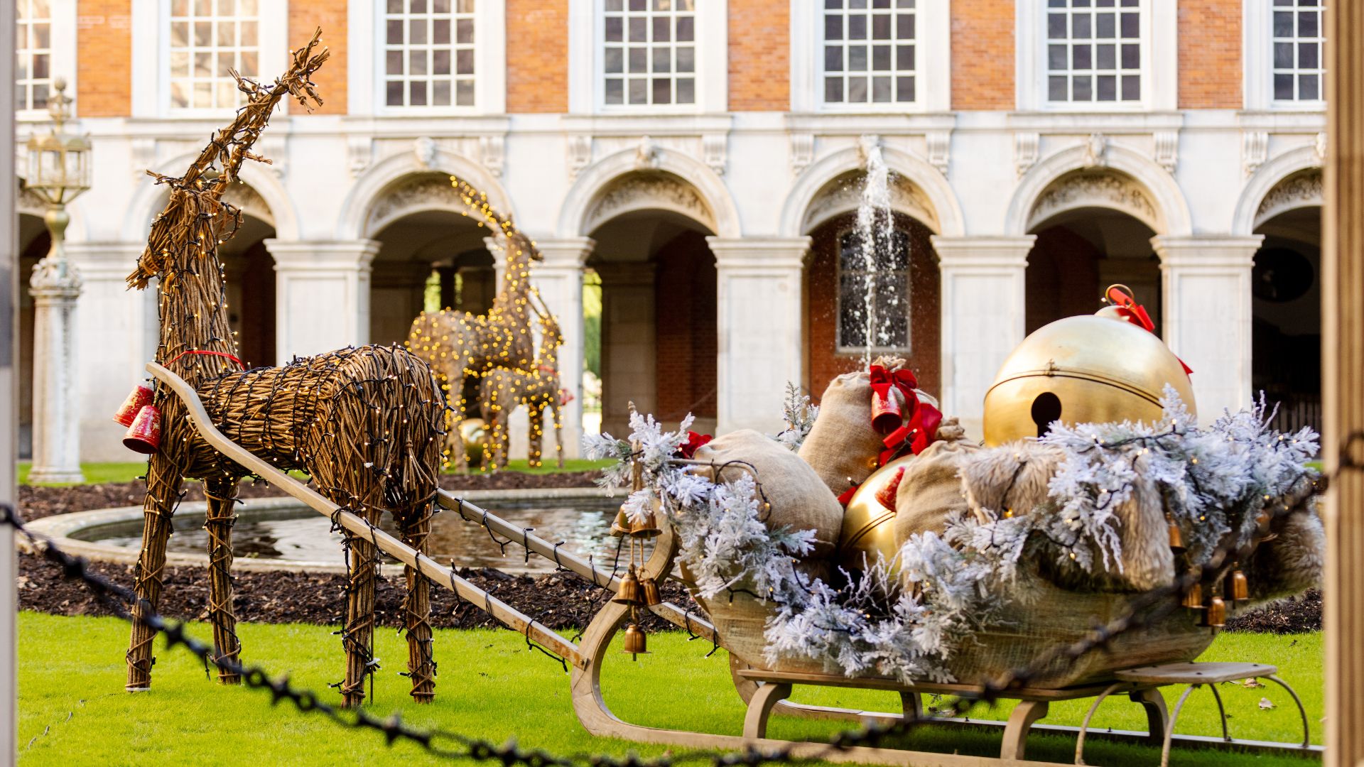 Visit Hampton Court Palace for spectacular Christmas decorations and events. Hampton Court Palace: © Historic Royal Palaces. Sled with Reindeer and a Christmas present bag and the exterior of Hampton Court Palace in the background.
