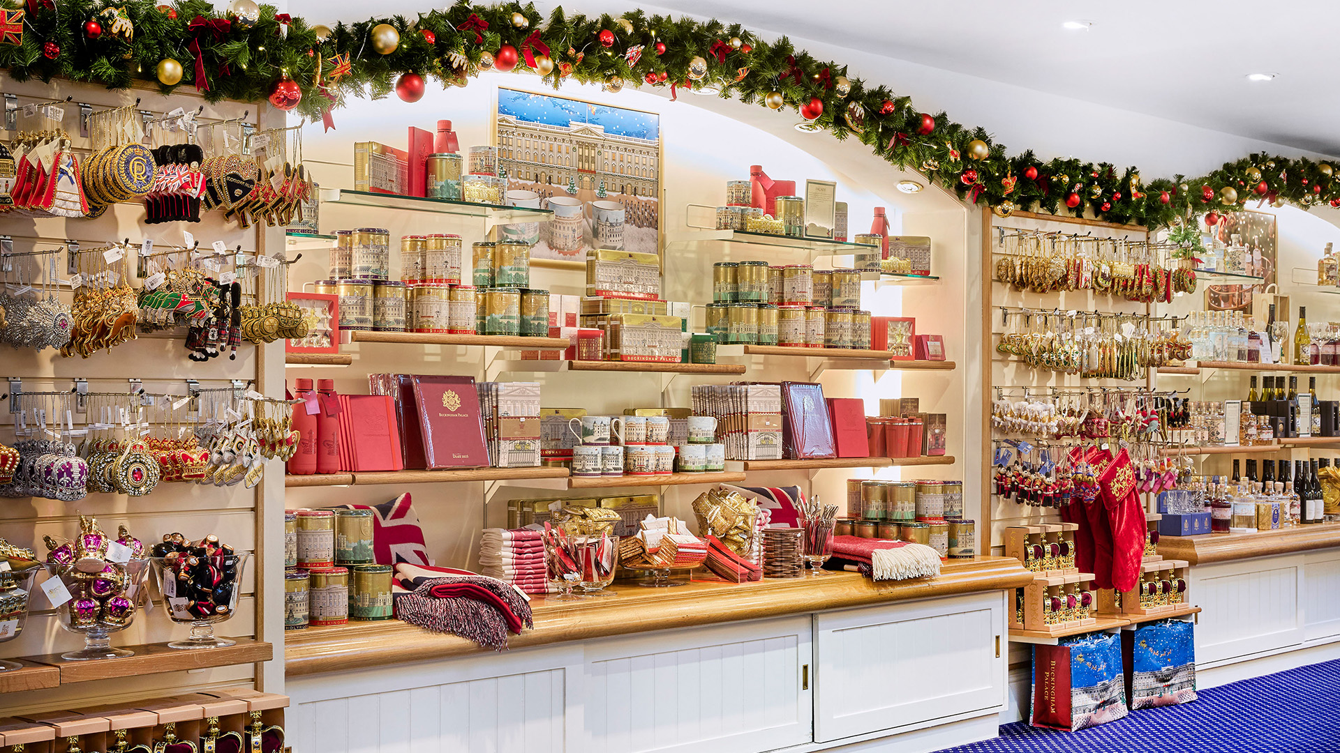 Visit Christmas pop-up shop at the Royal Mews © Royal Collection Enterprises Limited 2025 | Royal Collection Trust. A wall decorated with a Christmas garland and fitted with cupboards filled with gifts for Christmas