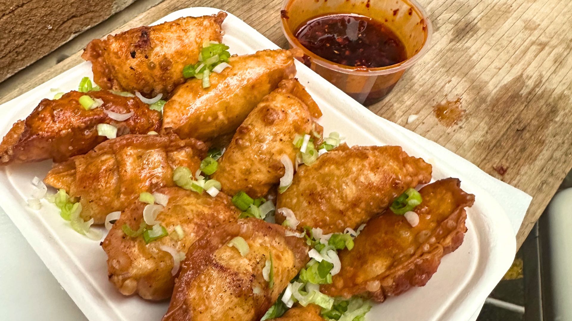 Mr Dumpling's chicken dumplings sprinkled with spring onions on a takeaway plate, plus a pot of dipping sauce, on a bench.