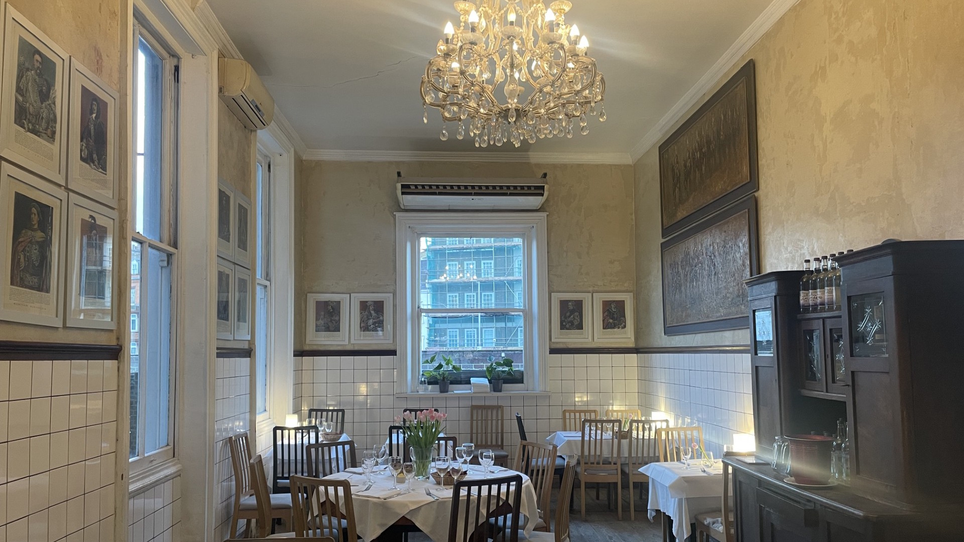 Tables with white tablecloths and traditional chairs in the dining room at Daquise, which also features white tiled walls and chandeliers.
