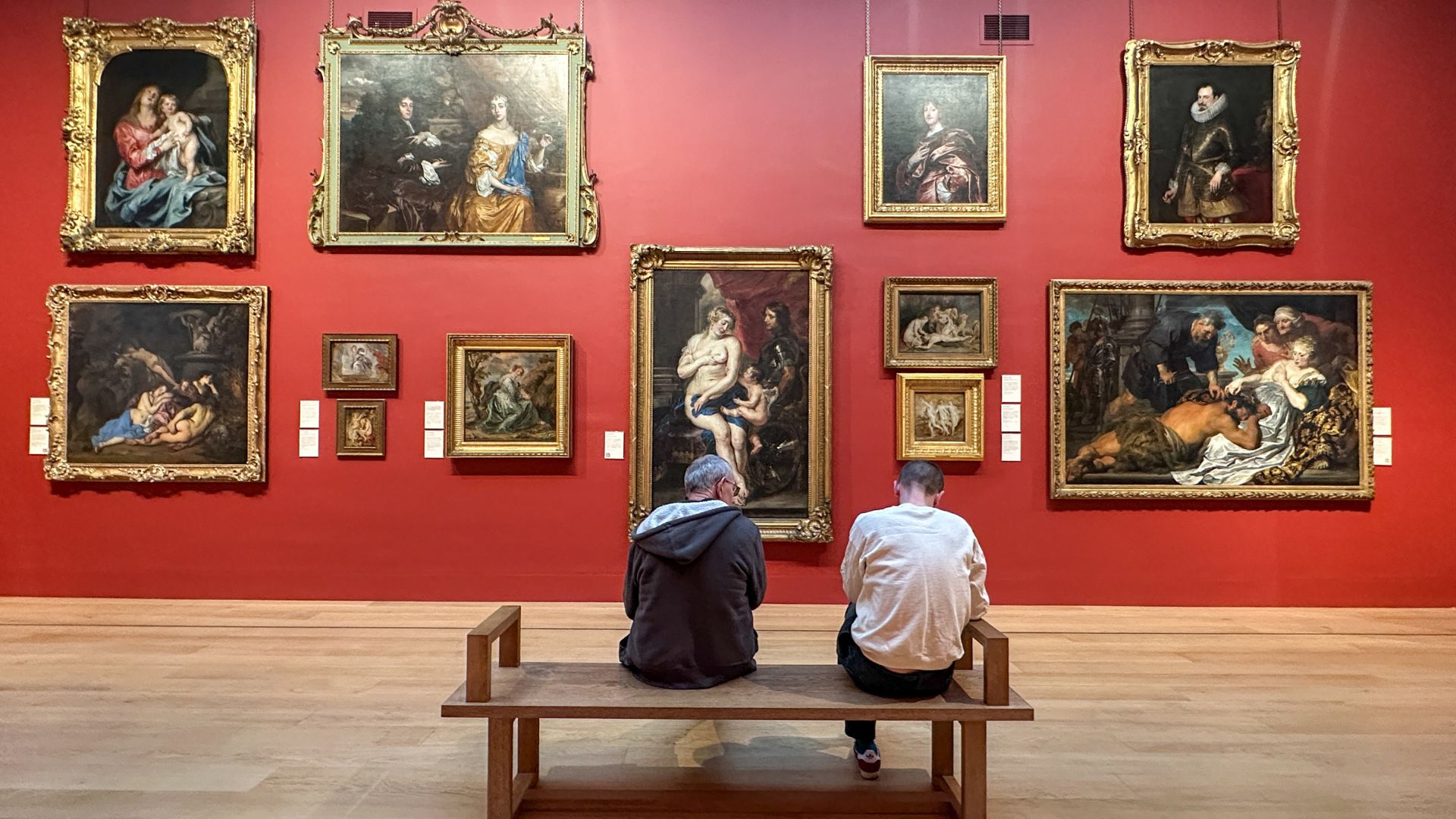 Two men sit on a brown bench looking at a red wall covered in paintings.