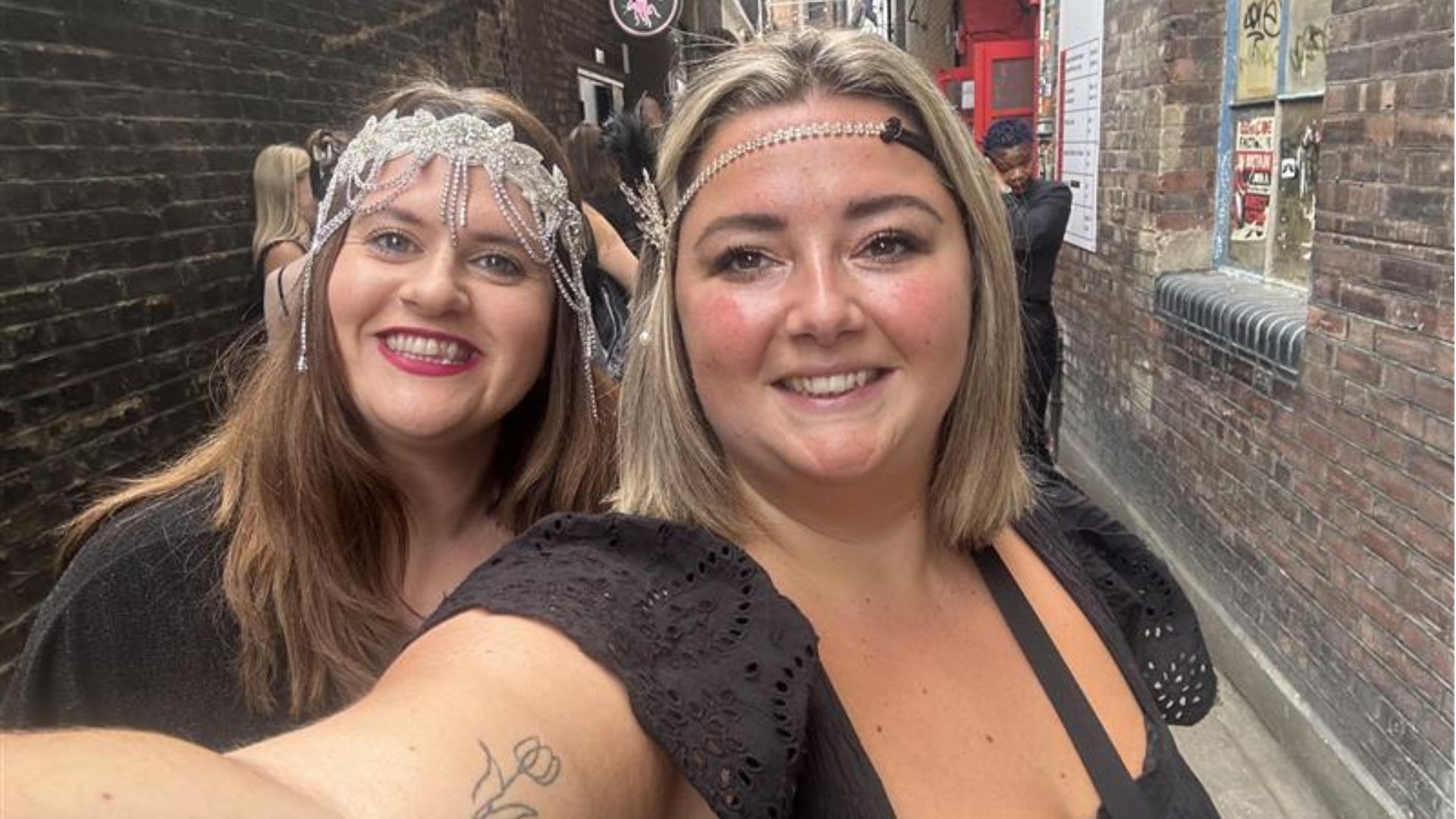 two women holding up a phone smiling and taking a selfie; both are wearing black dresses and wearing a 1930's-style feather and gem headband