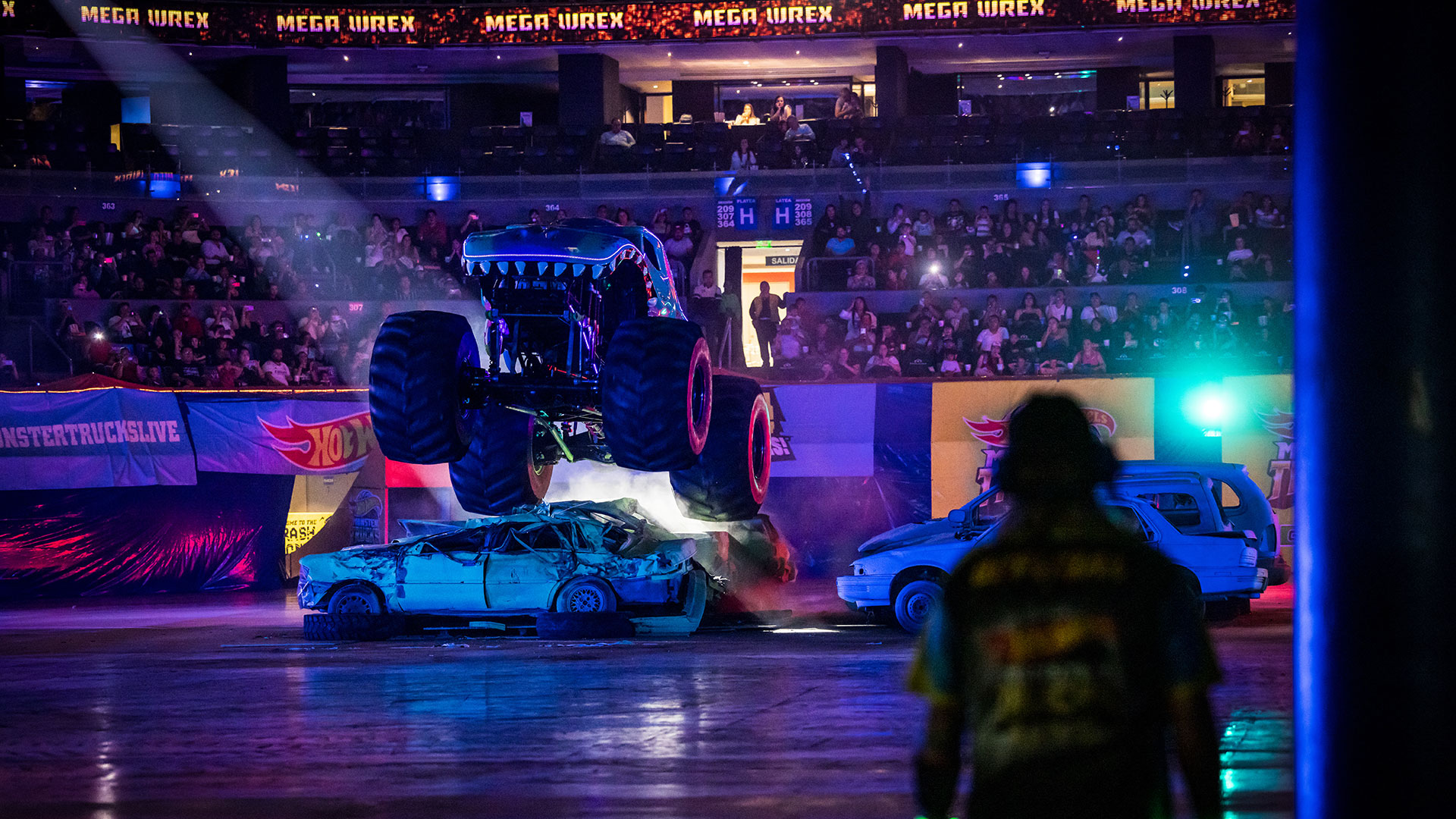 A Monster Truck jumps over a partly crushed car within an indoor arena.