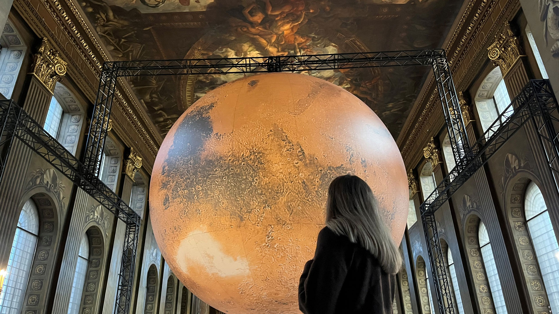 A lady looks up at the large Mars installation, a huge red sphere that looks like the planet, within the Painted Hall in the Old Royal Naval College.