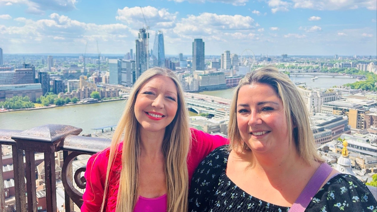 Two women pose in front of the camera at the top of St Paul's Cathedral, with a view of London's skyline behind, including the river Thames.