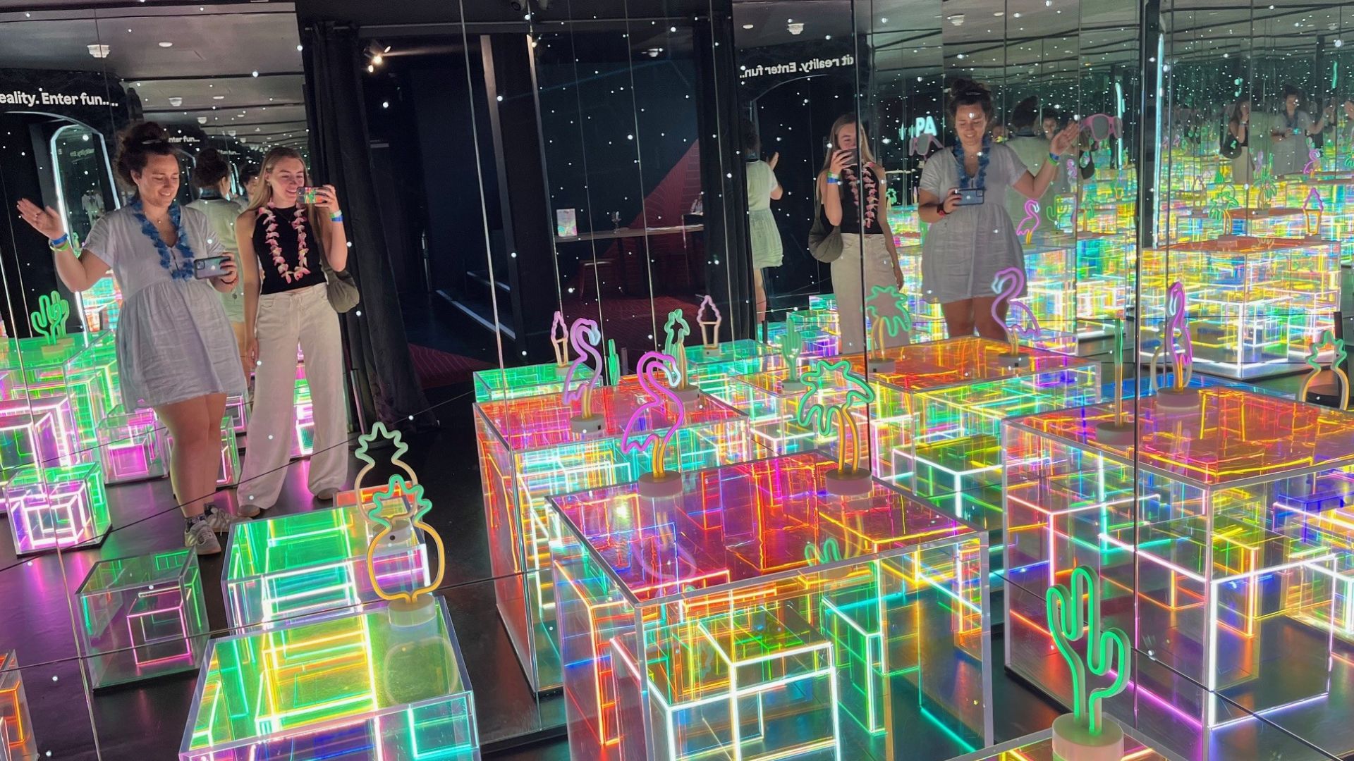See Paradox Museum transform into a Tropical Paradise this summer. © Visit London/Charlotte Pritchard. Two people standing in reflective mirror room with neon cactus and summer objects on the floor space.